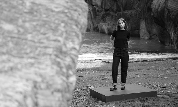 A woman stands confidently on a platform in front of coastal cliffs, wearing a black top and dark jeans. The black-and-white photo emphasises simplici